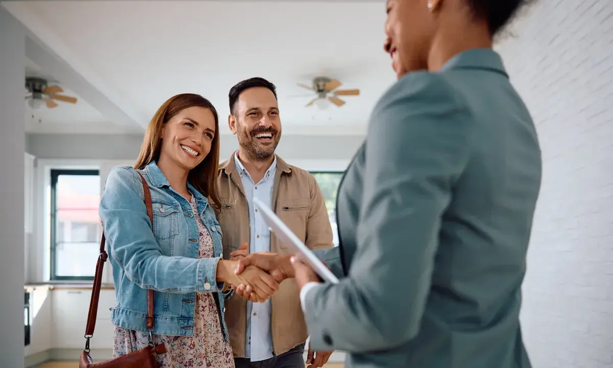 Un couple heureux serre la main d'une agente immobilière. Ils ont trouvé leur nouveau chez-soi, espérant que le loyer ne dépasse pas 3x leur salaire brut.