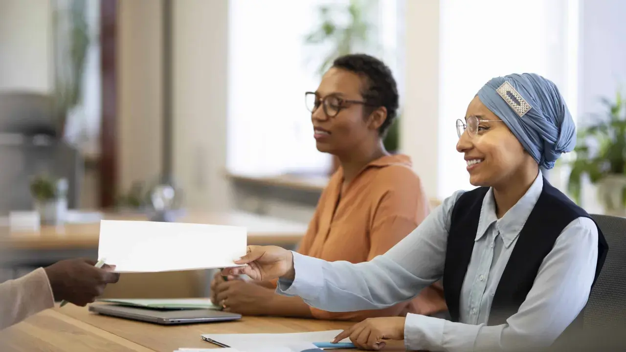 Une main tend un document blanc pour une vente en copropriété. Des femmes souriantes sont assises à une table, prêtes à examiner les documents obligatoires.