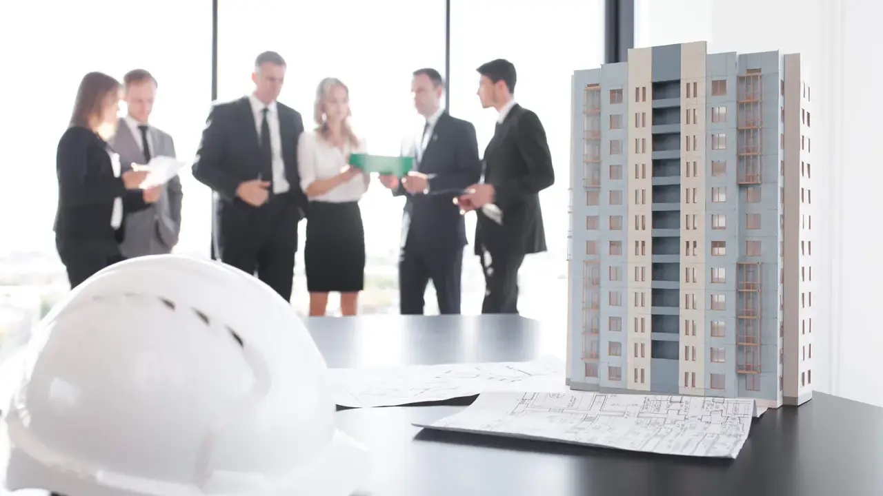 Maquette d'immeuble, plans et casque blanc sur une table. Des professionnels discutent, peut-être des travaux sur parties communes sans autorisation.