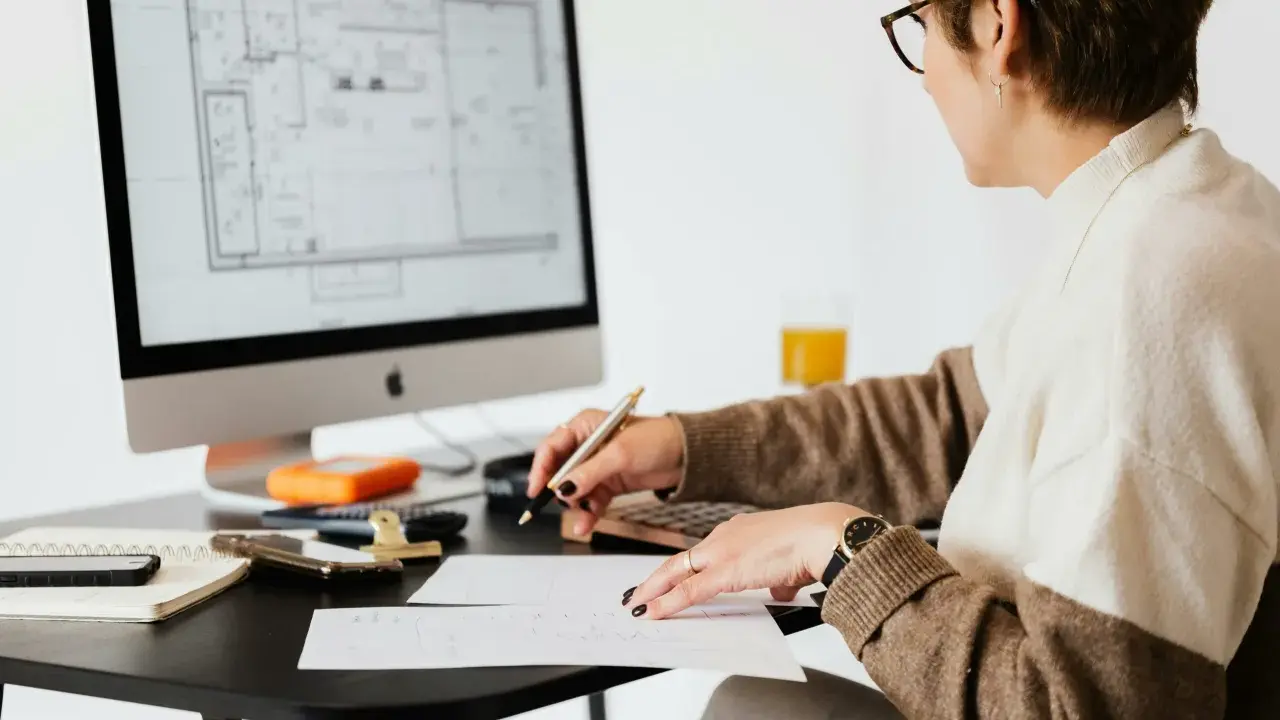 Une femme examine des plans sur son ordinateur, stylo à la main, calculatrice et notes à proximité. Elle est soulagée, car j'ai fini de payer mon crédit immobilier.