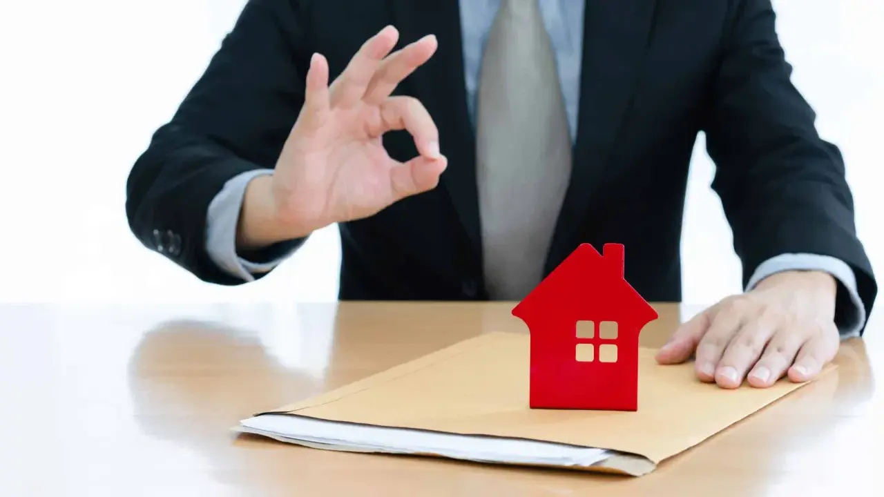 Un homme en costume fait un signe "OK" près d'une petite maison rouge sur une enveloppe. Un bon signe pour votre guide crédit immobilier.