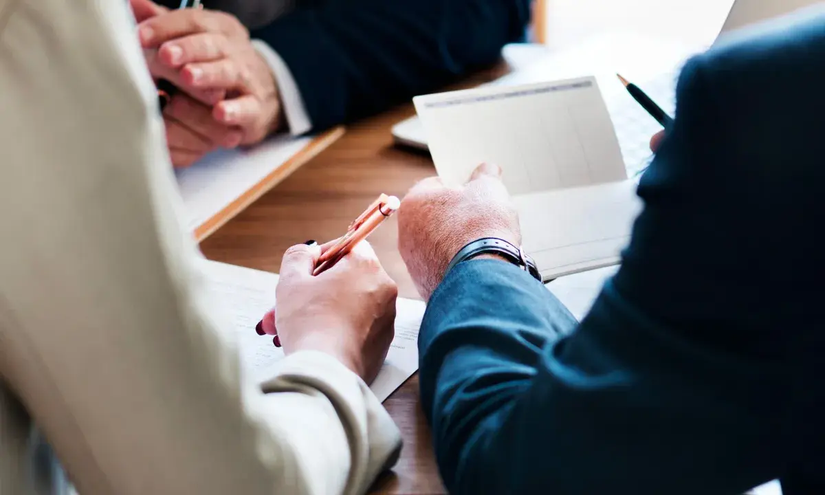 Trois mains se penchent sur un bureau en bois, stylo à la main, pour signer des documents. Le temps presse, il reste à vivre.
