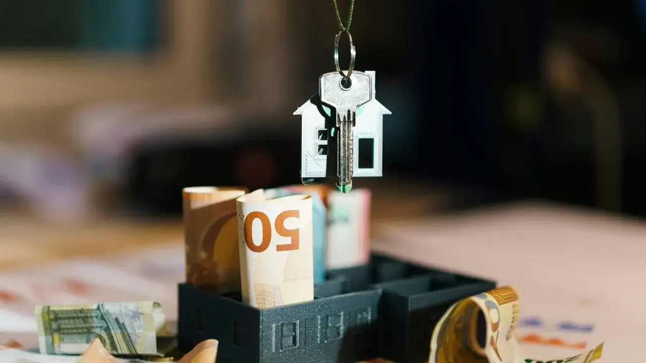 Clé d'une maison miniature suspendue au-dessus de billets d'euros roulés, symbolisant l'estimation taxe foncière.