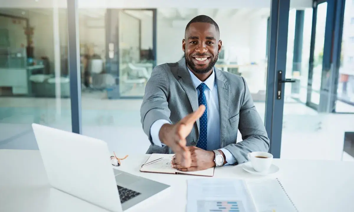Un homme d'affaires souriant tend la main pour une poignée de main, mais le vendeur ne veut plus vendre. Il est assis à un bureau avec un ordinateur portable et une tasse de café.