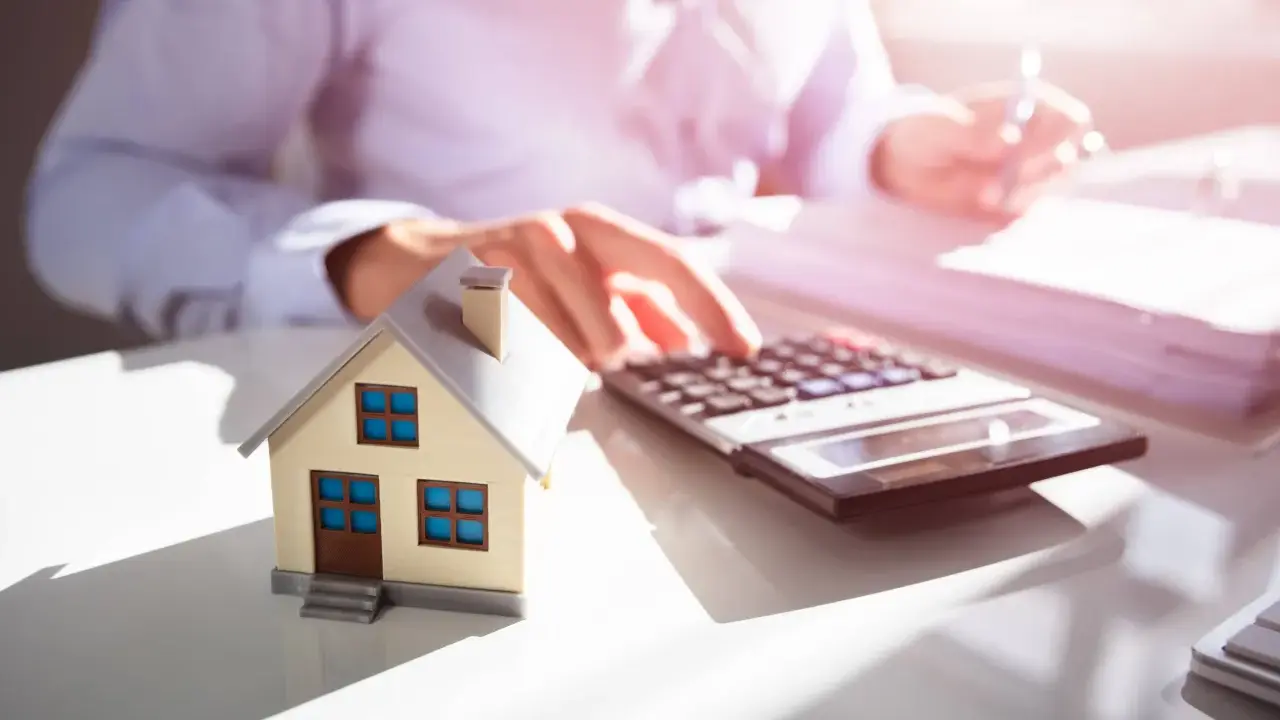 Maquette de maison sur un bureau, avec une calculatrice et des documents. Une personne calcule, probablement pour une estimation maison par notaire.