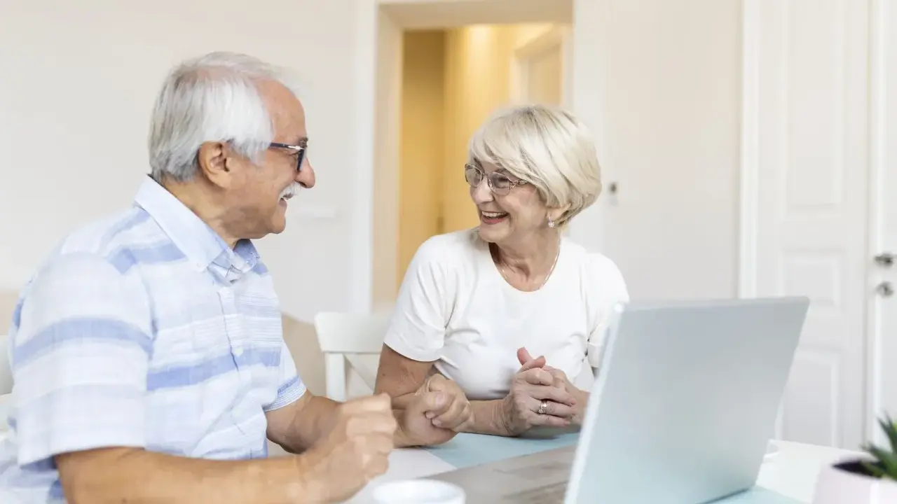 Couple âgé souriant devant un ordinateur, discutant peut-être de l'exonération taxe foncière 75 ans.