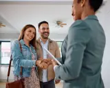 Un couple heureux serre la main d'une agente immobilière. Ils ont trouvé leur nouveau chez-soi, espérant que le loyer ne dépasse pas 3x leur salaire brut.