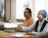 Une main tend un document blanc pour une vente en copropriété. Des femmes souriantes sont assises à une table, prêtes à examiner les documents obligatoires.