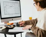 Une femme examine des plans sur son ordinateur, stylo à la main, calculatrice et notes à proximité. Elle est soulagée, car j'ai fini de payer mon crédit immobilier.