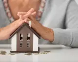 Simulation prêt immobilier après 60 ans : mains protégeant une maison miniature, avec des pièces sur la table.