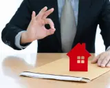 Un homme en costume fait un signe "OK" près d'une petite maison rouge sur une enveloppe. Un bon signe pour votre guide crédit immobilier.