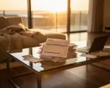 Pile de factures sur une table basse en verre, devant un canapé et de grandes fenêtres baignées de soleil. La scène évoque une résidence principale non habitée, peut-être en attente de gestion.
