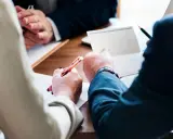 Trois mains se penchent sur un bureau en bois, stylo à la main, pour signer des documents. Le temps presse, il reste à vivre.
