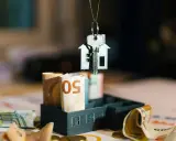 Clé d'une maison miniature suspendue au-dessus de billets d'euros roulés, symbolisant l'estimation taxe foncière.
