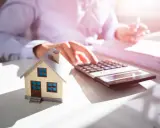 Maquette de maison sur un bureau, avec une calculatrice et des documents. Une personne calcule, probablement pour une estimation maison par notaire.