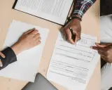 Des mains signent un compromis de vente sur un bureau en bois, entourées de documents.