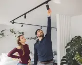 Un homme ajuste un spot sur un rail noir, sous une belle hauteur sous plafond. Une femme le regarde, curieuse.