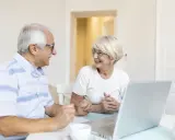 Couple âgé souriant devant un ordinateur, discutant peut-être de l'exonération taxe foncière 75 ans.