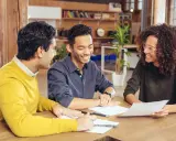 Trois personnes discutent autour d'une table, examinant des documents. Ils semblent planifier leur projet et se demandent combien emprunter avec 2500 euros par mois.