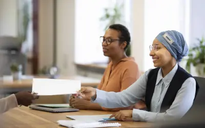Une main tend un document blanc pour une vente en copropriété. Des femmes souriantes sont assises à une table, prêtes à examiner les documents obligatoires.