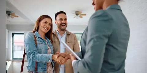 Un couple heureux serre la main d'une agente immobilière. Ils ont trouvé leur nouveau chez-soi, espérant que le loyer ne dépasse pas 3x leur salaire brut.