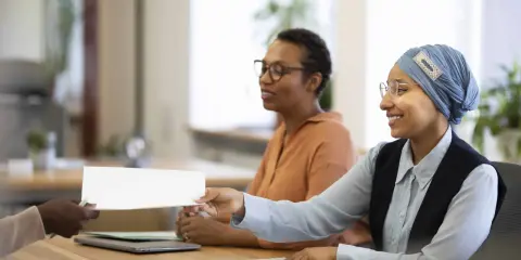 Une main tend un document blanc pour une vente en copropriété. Des femmes souriantes sont assises à une table, prêtes à examiner les documents obligatoires.