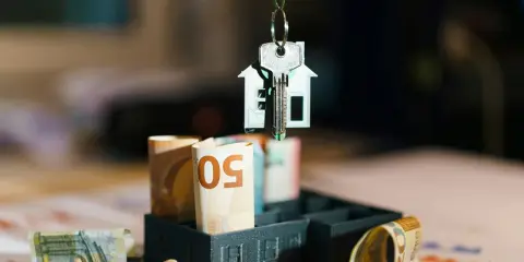 Clé d'une maison miniature suspendue au-dessus de billets d'euros roulés, symbolisant l'estimation taxe foncière.