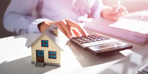 Maquette de maison sur un bureau, avec une calculatrice et des documents. Une personne calcule, probablement pour une estimation maison par notaire.