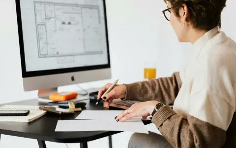 Une femme examine des plans sur son ordinateur, stylo à la main, calculatrice et notes à proximité. Elle est soulagée, car j'ai fini de payer mon crédit immobilier.