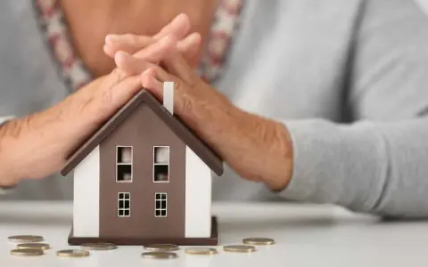 Simulation prêt immobilier après 60 ans : mains protégeant une maison miniature, avec des pièces sur la table.