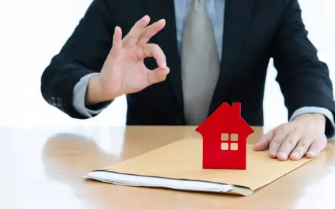 Un homme en costume fait un signe "OK" près d'une petite maison rouge sur une enveloppe. Un bon signe pour votre guide crédit immobilier.