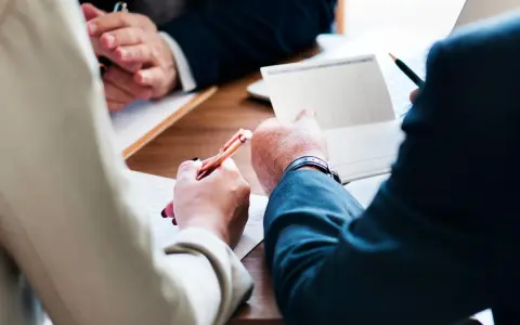 Trois mains se penchent sur un bureau en bois, stylo à la main, pour signer des documents. Le temps presse, il reste à vivre.