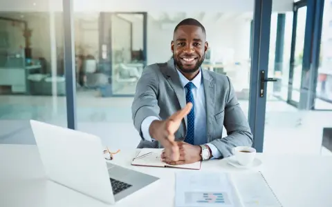 Un homme d'affaires souriant tend la main pour une poignée de main, mais le vendeur ne veut plus vendre. Il est assis à un bureau avec un ordinateur portable et une tasse de café.