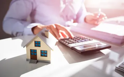 Maquette de maison sur un bureau, avec une calculatrice et des documents. Une personne calcule, probablement pour une estimation maison par notaire.