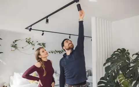 Un homme ajuste un spot sur un rail noir, sous une belle hauteur sous plafond. Une femme le regarde, curieuse.