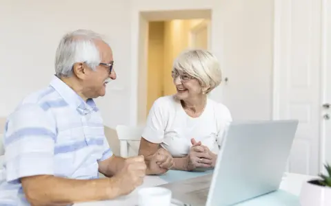 Couple âgé souriant devant un ordinateur, discutant peut-être de l'exonération taxe foncière 75 ans.