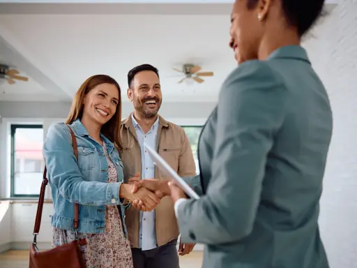 Un couple heureux serre la main d'une agente immobilière. Ils ont trouvé leur nouveau chez-soi, espérant que le loyer ne dépasse pas 3x leur salaire brut.
