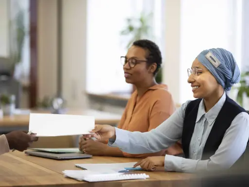 Une main tend un document blanc pour une vente en copropriété. Des femmes souriantes sont assises à une table, prêtes à examiner les documents obligatoires.