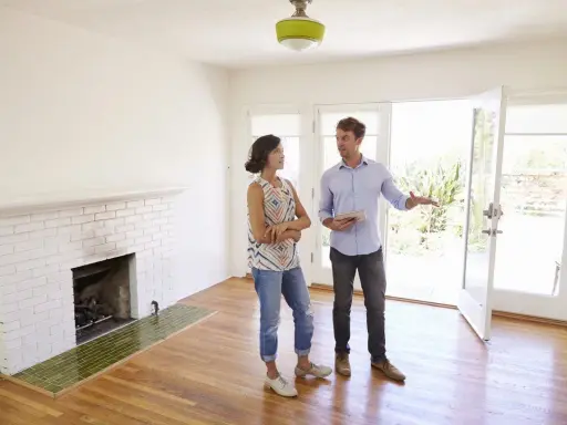 Un agent immobilier présente un appartement ou une maison à une cliente potentielle. La pièce est lumineuse, avec une cheminée en briques blanches et un sol en bois.
