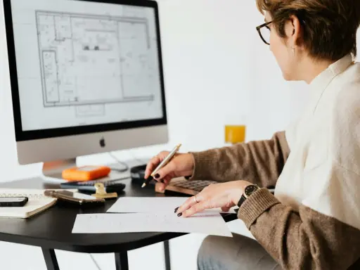 Une femme examine des plans sur son ordinateur, stylo à la main, calculatrice et notes à proximité. Elle est soulagée, car j'ai fini de payer mon crédit immobilier.