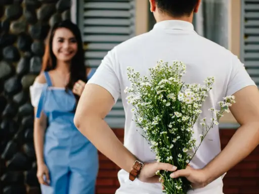 Un homme cache un bouquet de fleurs derrière son dos, prêt à surprendre sa compagne. Un couple non marié dans un lieu extérieur.