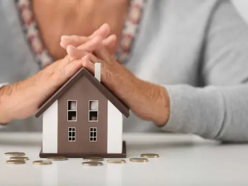 Simulation prêt immobilier après 60 ans : mains protégeant une maison miniature, avec des pièces sur la table.