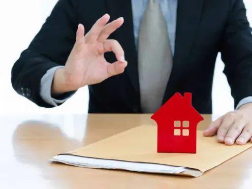 Un homme en costume fait un signe "OK" près d'une petite maison rouge sur une enveloppe. Un bon signe pour votre guide crédit immobilier.
