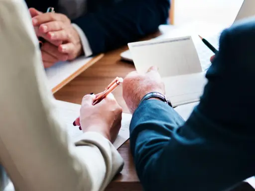 Trois mains se penchent sur un bureau en bois, stylo à la main, pour signer des documents. Le temps presse, il reste à vivre.