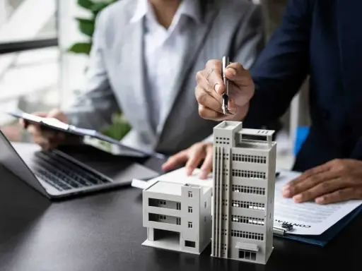 Deux personnes discutent de modèles de bâtiments et de documents, possiblement pour un projet immobilier ou un régime fiscal immobilier.