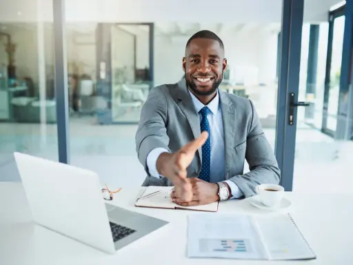 Un homme d'affaires souriant tend la main pour une poignée de main, mais le vendeur ne veut plus vendre. Il est assis à un bureau avec un ordinateur portable et une tasse de café.