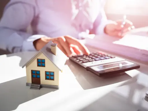 Maquette de maison sur un bureau, avec une calculatrice et des documents. Une personne calcule, probablement pour une estimation maison par notaire.