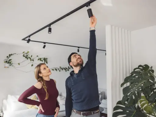 Un homme ajuste un spot sur un rail noir, sous une belle hauteur sous plafond. Une femme le regarde, curieuse.