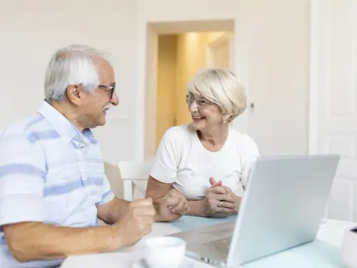 Couple âgé souriant devant un ordinateur, discutant peut-être de l'exonération taxe foncière 75 ans.