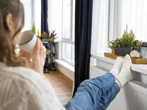 Une personne boit une tasse chaude, les pieds sur un radiateur, profitant de la chaleur. Un bon DPE chauffage électrique pour un confort optimal.